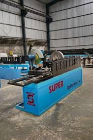 The image shows an industrial **roll forming machine** by **Super Rollforming**, designed for shaping metal sheets into specific profiles.  The machine is painted in a bright blue color with the brand name clearly visible on its body. It features a long production line with multiple roller stations that gradually form the metal as it passes through. At one end, a cutting or pressing section is visible, which is used to shape or cut the material into the required design.  The setup is placed inside a spacious factory with a high roof and metal walls, indicating an industrial environment. The machine appears robust, efficient, and suitable for continuous, large-scale manufacturing operations.