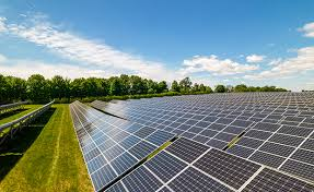The image shows a large solar power installation set in an open outdoor area. Rows of solar panels are arranged in long, neat lines across a wide field. The panels are tilted at an angle to capture maximum sunlight, which helps generate electricity efficiently.